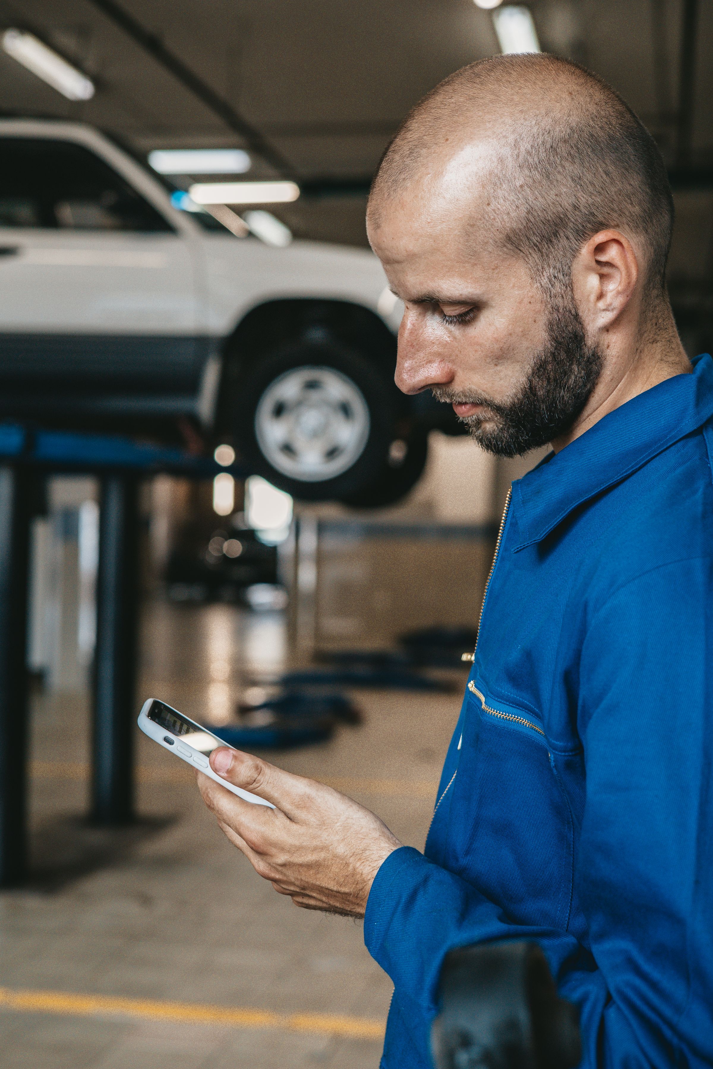 A mechanic in blue overalls reading an enquiry on his phone, a car raised on the lift behind him